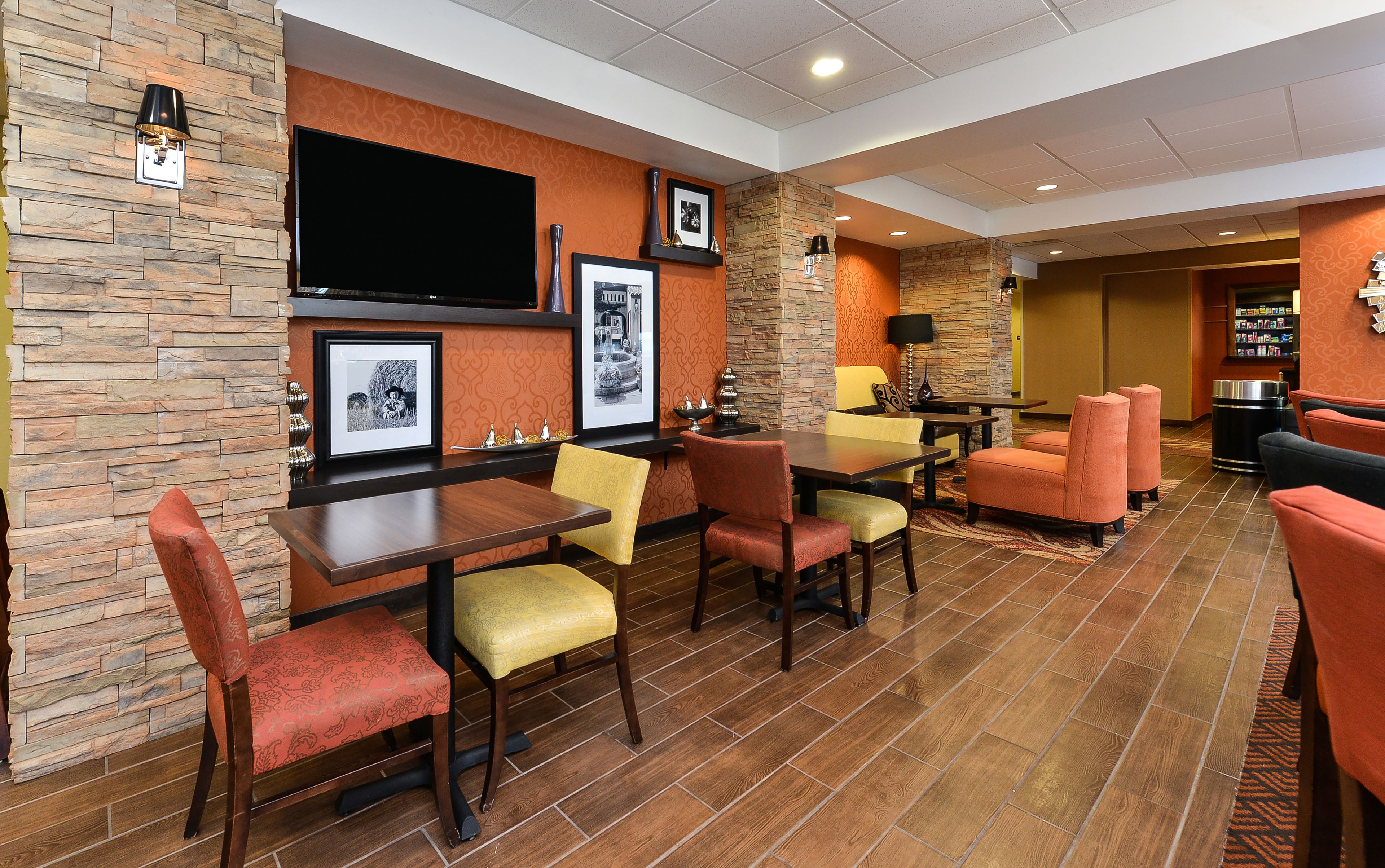 A hotel lobby with a stone wall, a flat-screen TV, framed pictures, and a seating area with yellow and red chairs.
