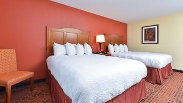 A hotel room with two beds, a wooden headboard, a bedside table with a lamp, a framed picture on the wall, and a patterned chair.