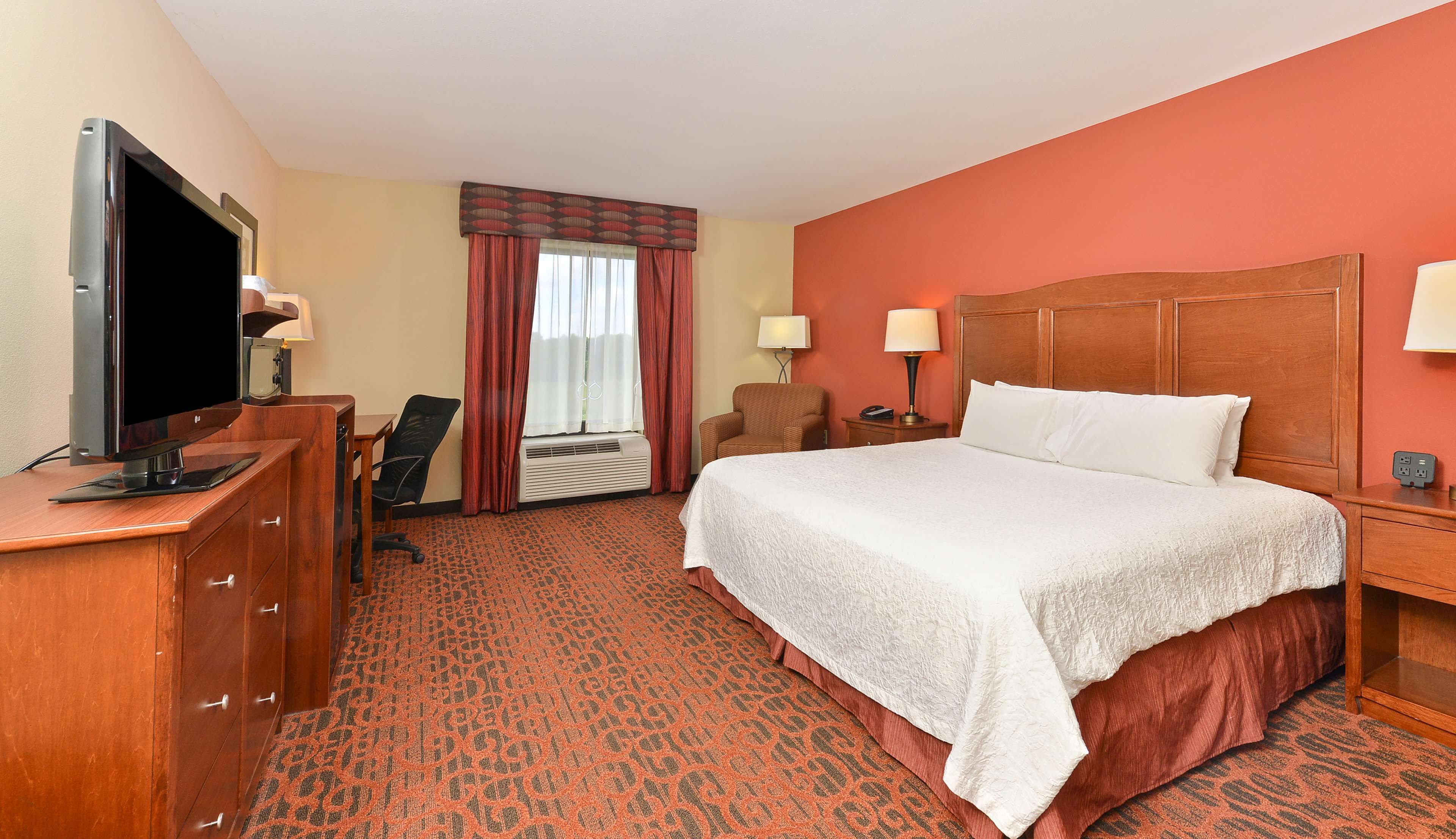 A hotel room with a large bed, a desk with a flat-screen TV, a chair, a window with curtains, and a carpeted floor.
