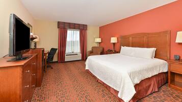 A hotel room with a large bed, a desk with a flat-screen TV, a chair, a window with curtains, and a carpeted floor.