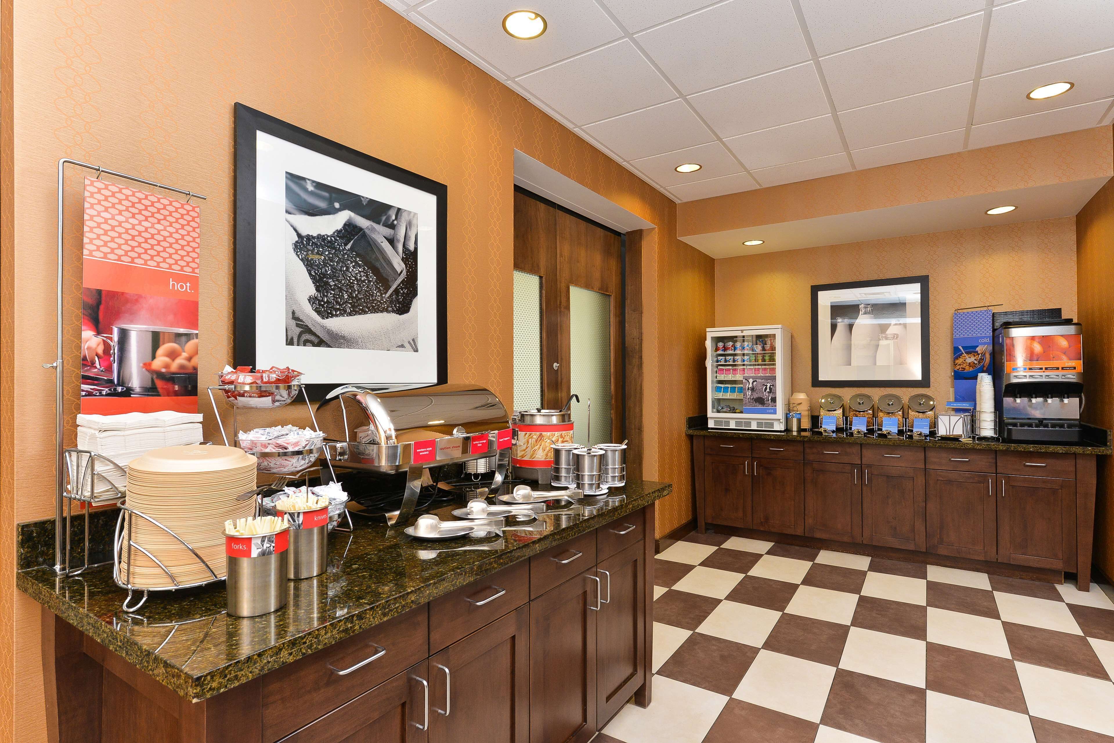 A hotel breakfast area with a buffet setup, including various food containers, a coffee machine, and a refrigerator.