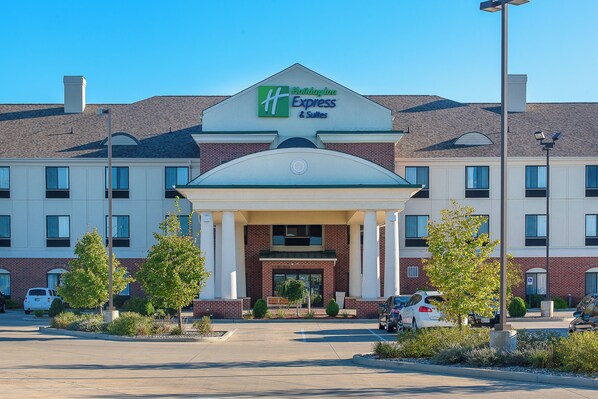 Un hôtel Holiday Inn Express & Suites avec une façade en briques, une entrée centrale dotée d’un porche abrité et un parking où sont stationnées plusieurs voitures.