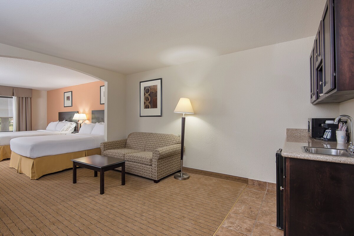 Une chambre d’hôtel comprenant un lit, un canapé, une petite table, une lampe, un tableau au mur et un coin cuisine.