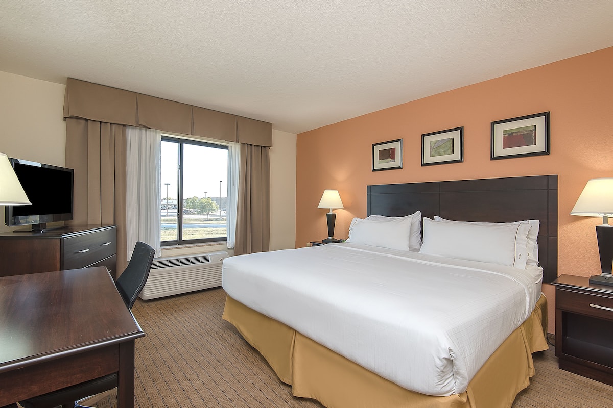 A hotel room with a large bed, a desk with a television, a window with curtains, and two wall-mounted lamps.
