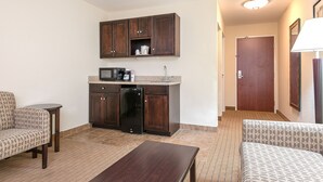 A compact hotel room with a kitchenette, two patterned sofas, a small wooden table, and a door leading to another room. - Holiday Inn Express & Suites Lafayette East by IHG (Lafayette)