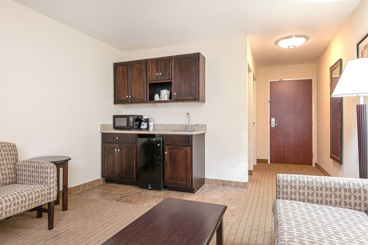 A compact hotel room with a kitchenette, two patterned sofas, a small wooden table, and a door leading to another room.