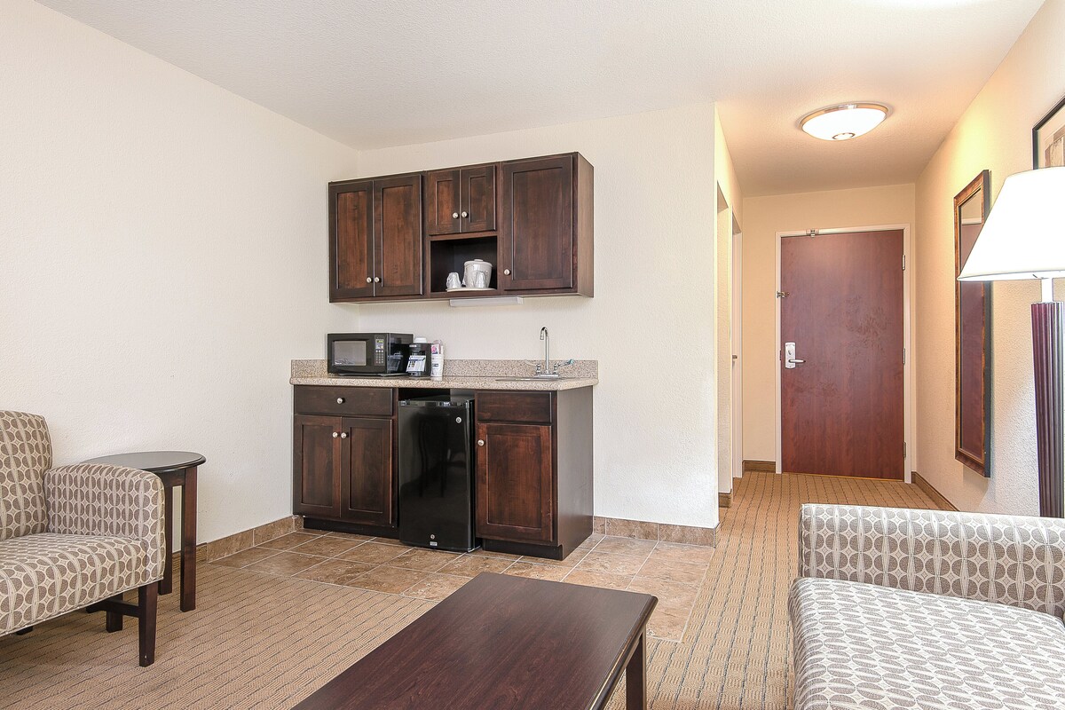 Une chambre d’hôtel compacte dotée d’une kitchenette, d’un coin salon avec deux fauteuils, d’une petite table et d’une porte donnant sur une autre pièce.