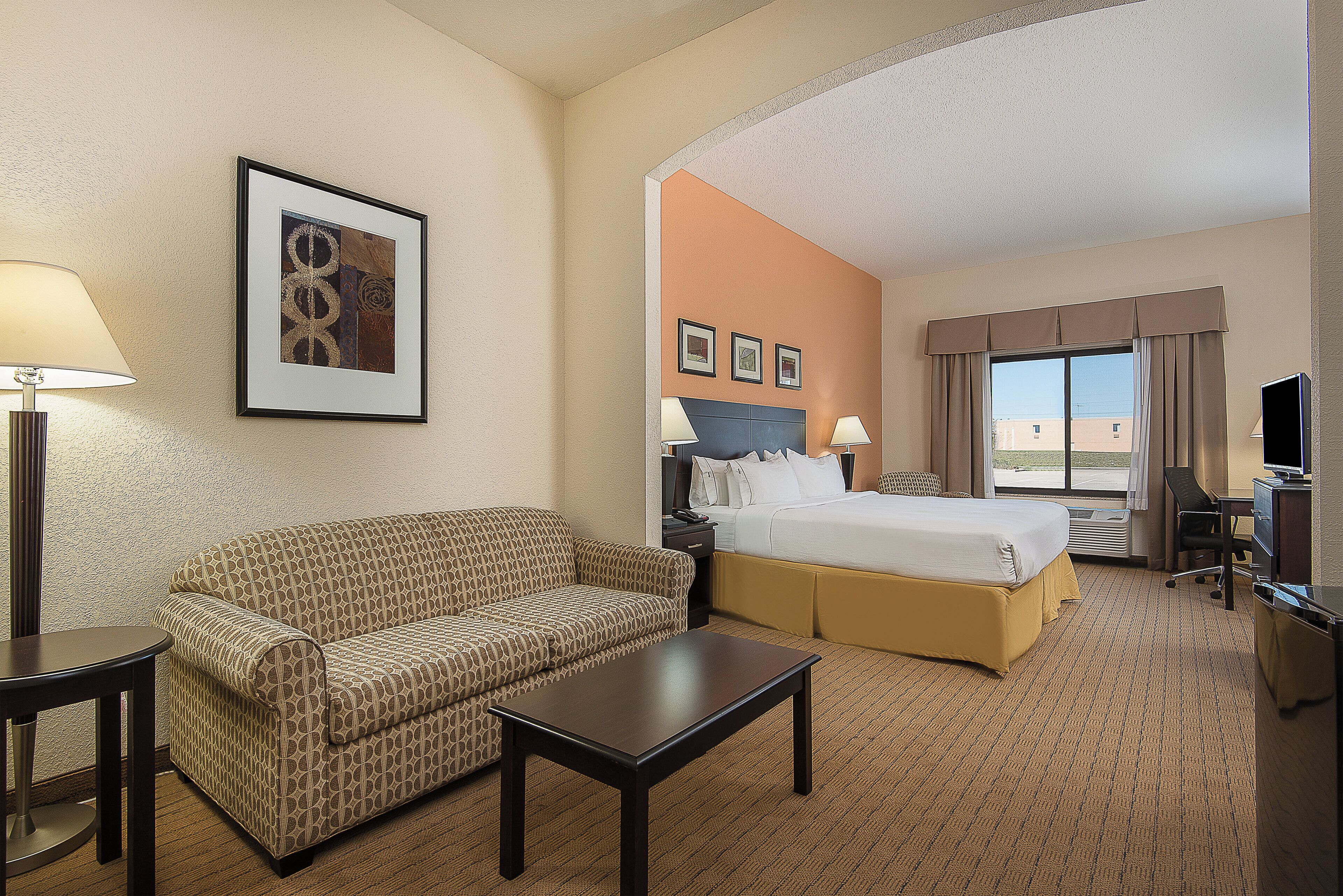 A hotel room with a bed, a sofa, a desk, a chair, a lamp, and a picture on the wall.