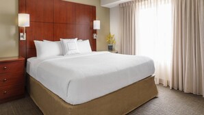 Quarto de hotel com uma cama grande, cabeceira de madeira, duas luminárias de cabeceira, mesa de apoio e uma janela com cortinas.