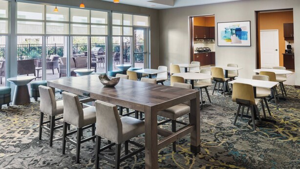 Quarto de hotel com uma grande mesa de jantar, várias cadeiras e piso coberto por carpete. Janelas amplas oferecem luz natural e vista para o exterior.