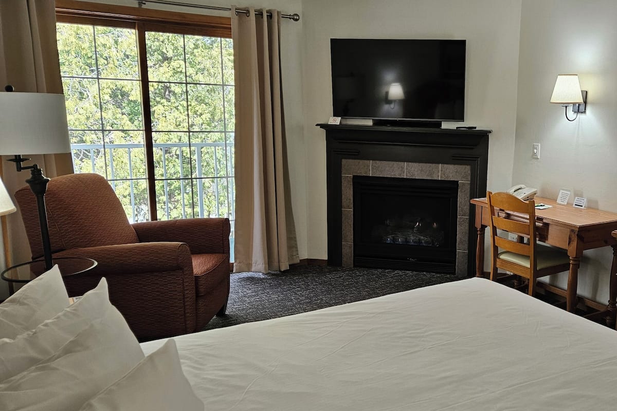 A hotel room with a bed, a desk, a chair, a fireplace, a television, and a large window with curtains.