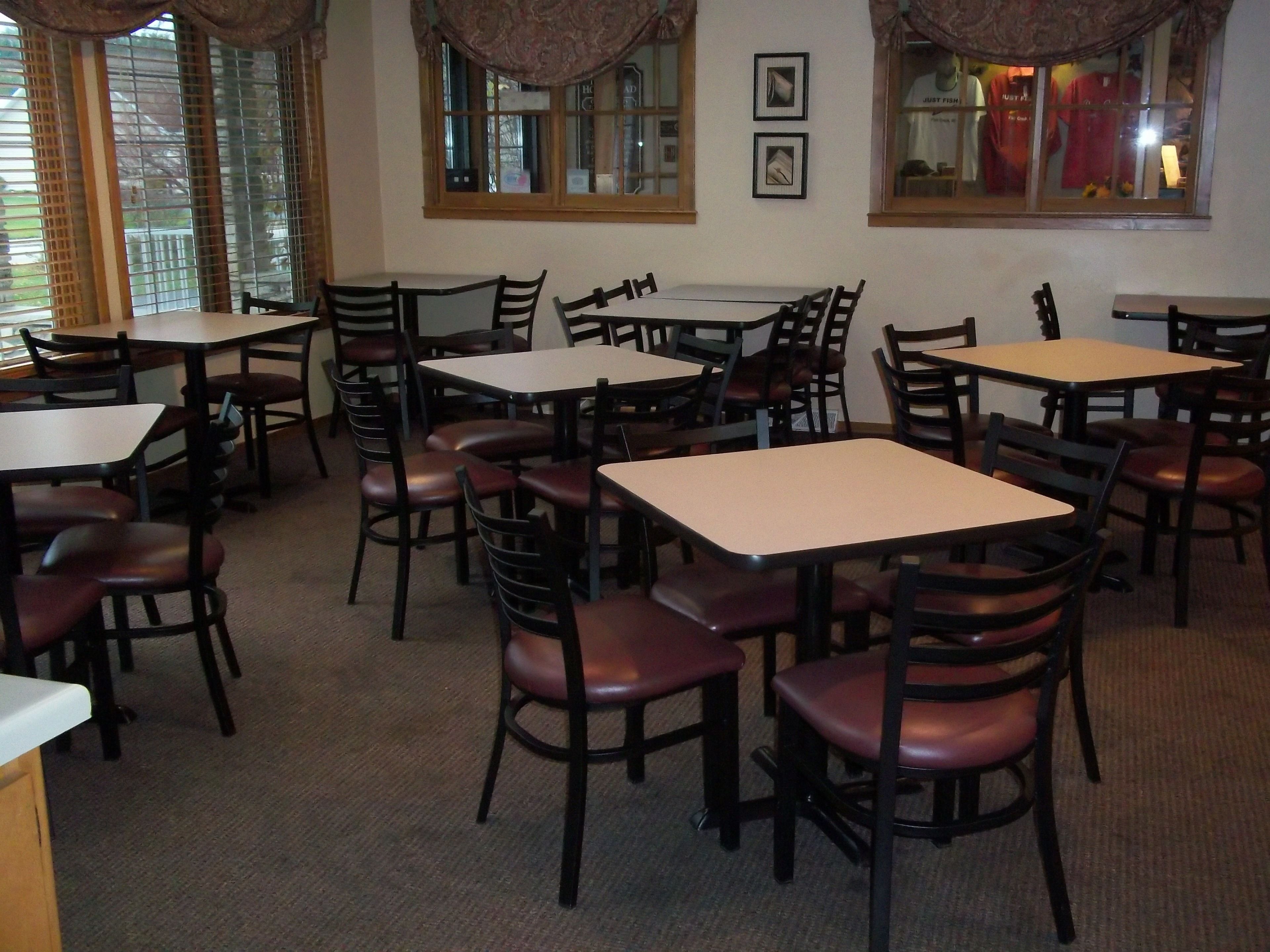 A dining area with multiple tables and chairs, large windows with curtains, and framed pictures on the wall.