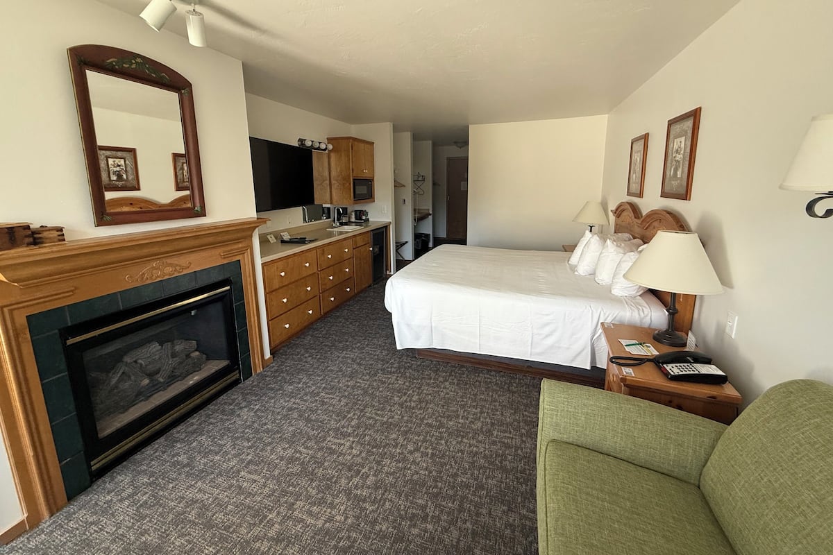 A hotel room with a fireplace, a bed with white bedding, a green armchair, a desk with a lamp, and a small table with a phone and papers.