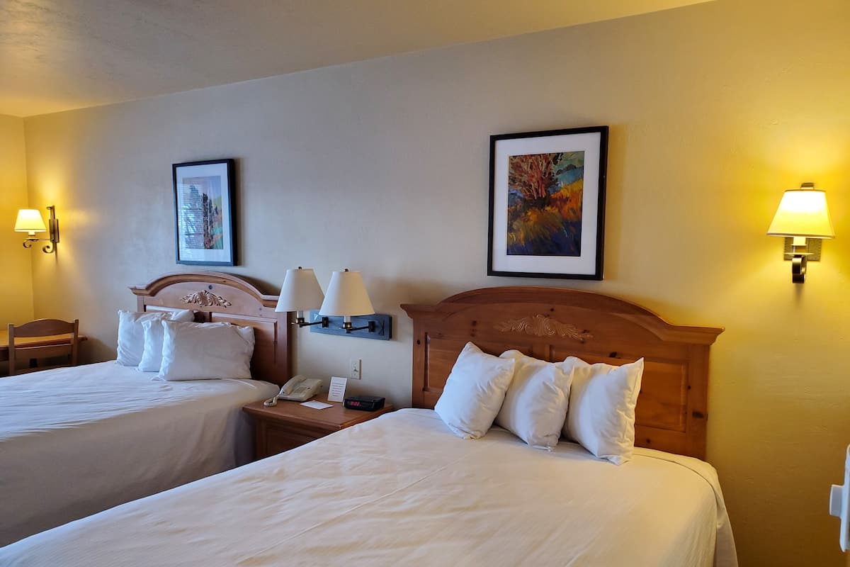 A hotel room with two beds, a wooden headboard, bedside lamps, a nightstand with a phone and a lamp, and framed artwork on the walls.