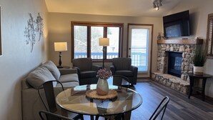 A living room with a glass table, two patterned armchairs, a sofa, a fireplace, and a TV mounted on the wall. - Homestead Suites (Fish Creek)