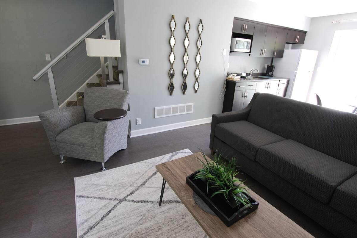 A modern living room with a grey sofa, a grey armchair, a wooden coffee table with a plant, a staircase, and a kitchen area with dark cabinets and a white refrigerator.