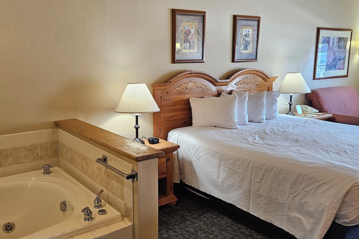 A hotel room with a large bed, a wooden headboard, two bedside lamps, a red armchair, and a bathtub.