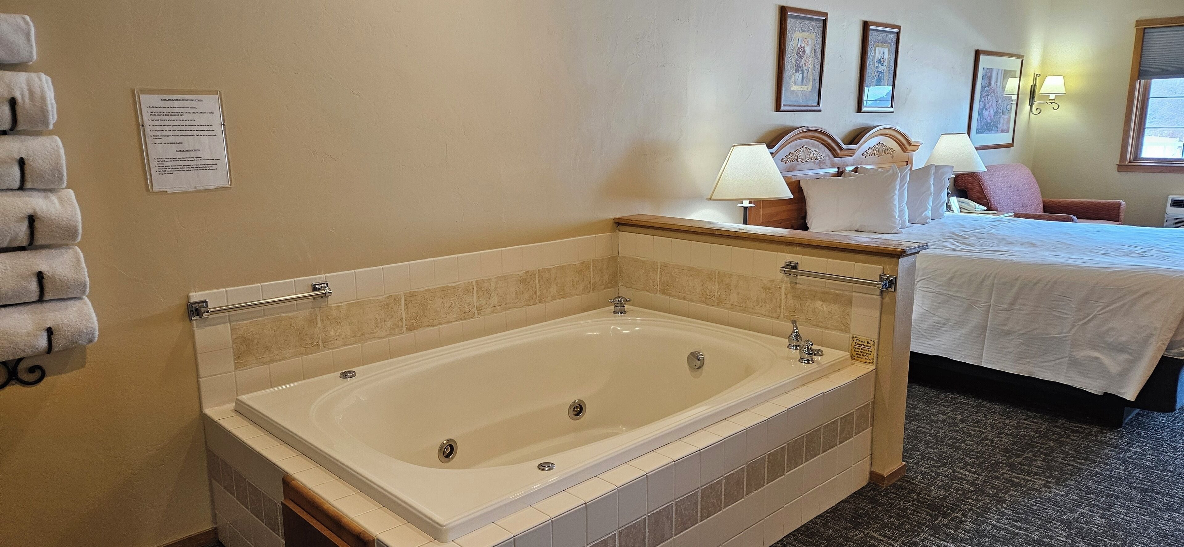 A hotel room with a large bathtub, a bed with white linens, a chair, and a wall-mounted lamp.