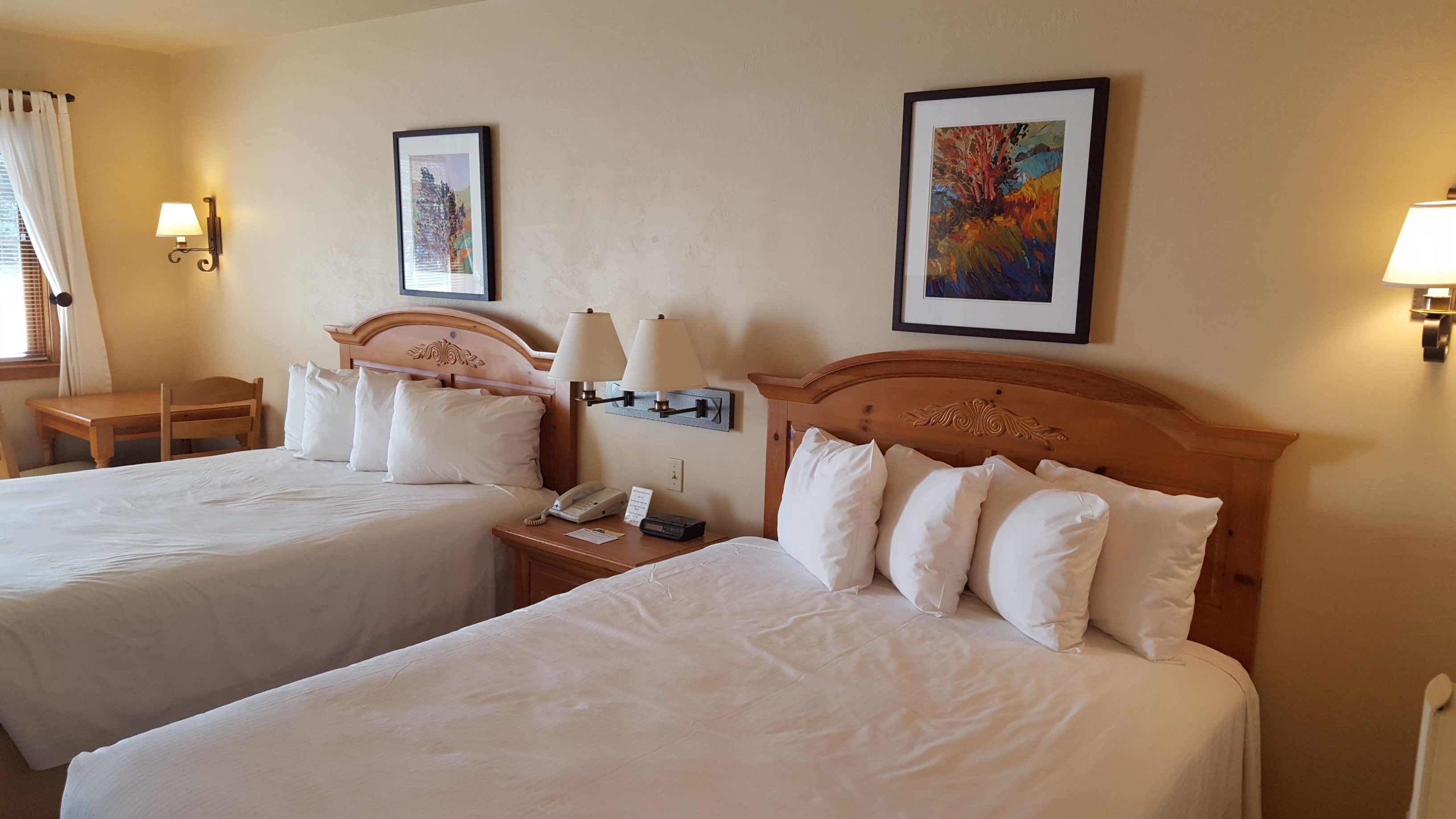 A hotel room with two beds, a desk, a chair, a nightstand with a phone and lamp, and two framed pictures on the wall.