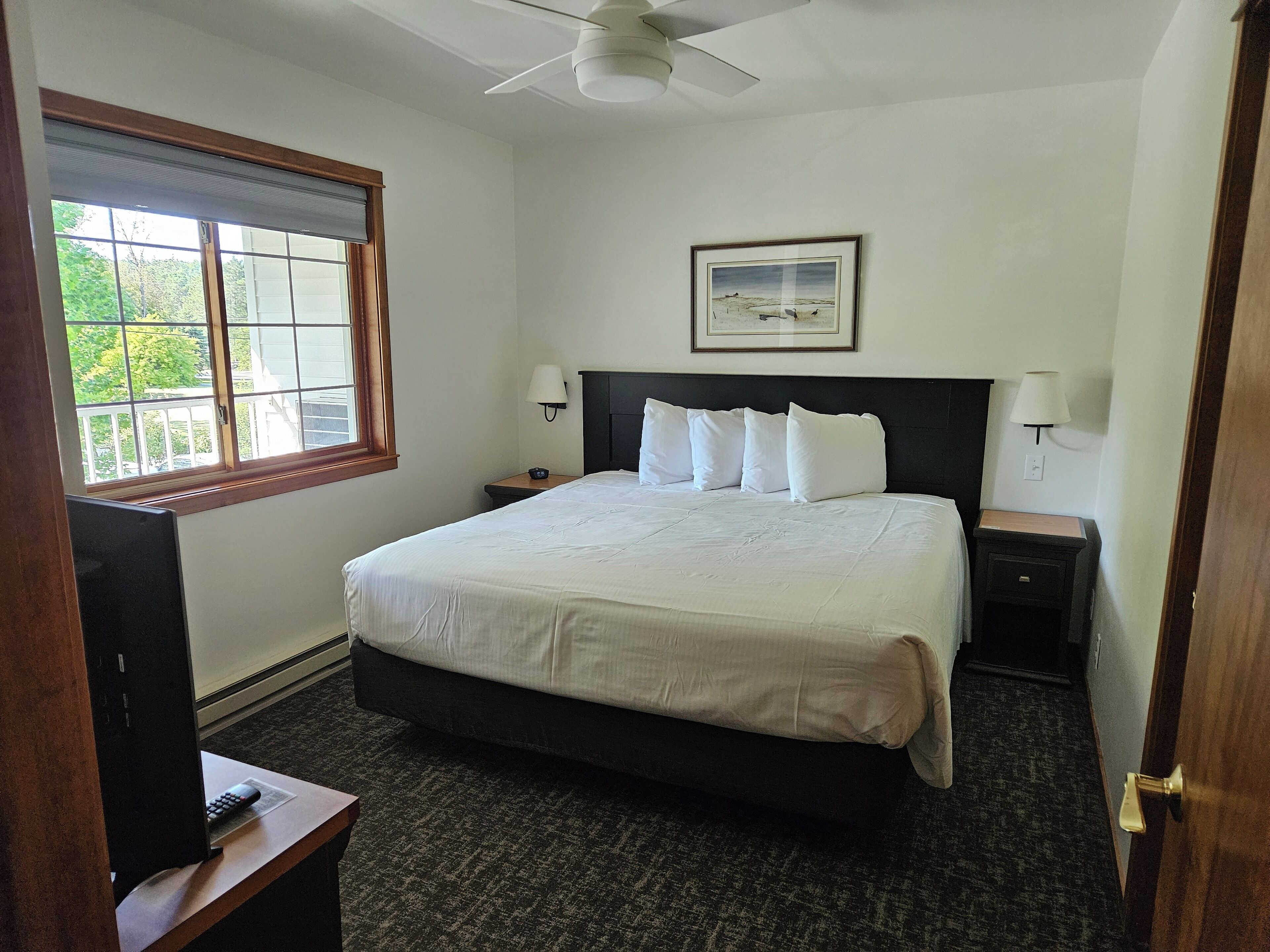 A neatly arranged bedroom with a bed, two bedside tables, a ceiling fan, a window with blinds, and a framed picture on the wall.