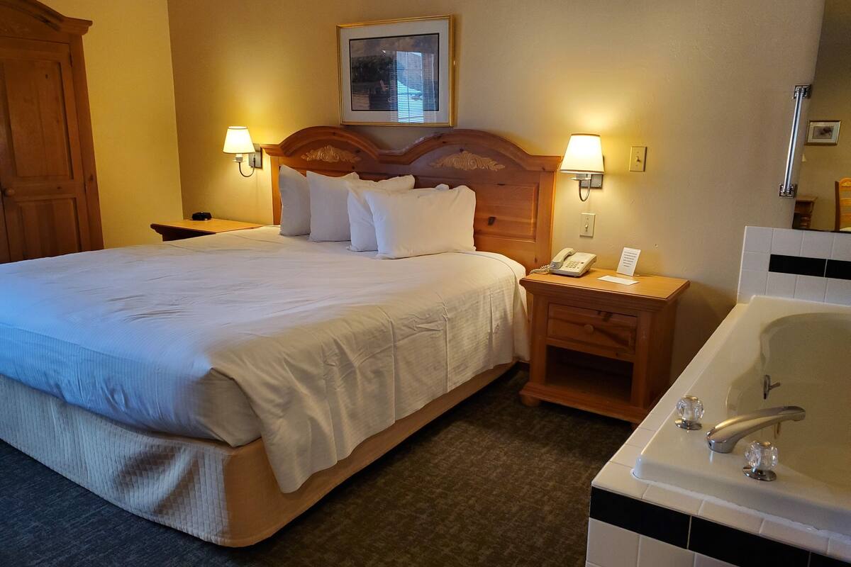 A hotel room with a large bed, two bedside lamps, a wooden nightstand with a phone, and a separate bathroom with a bathtub.