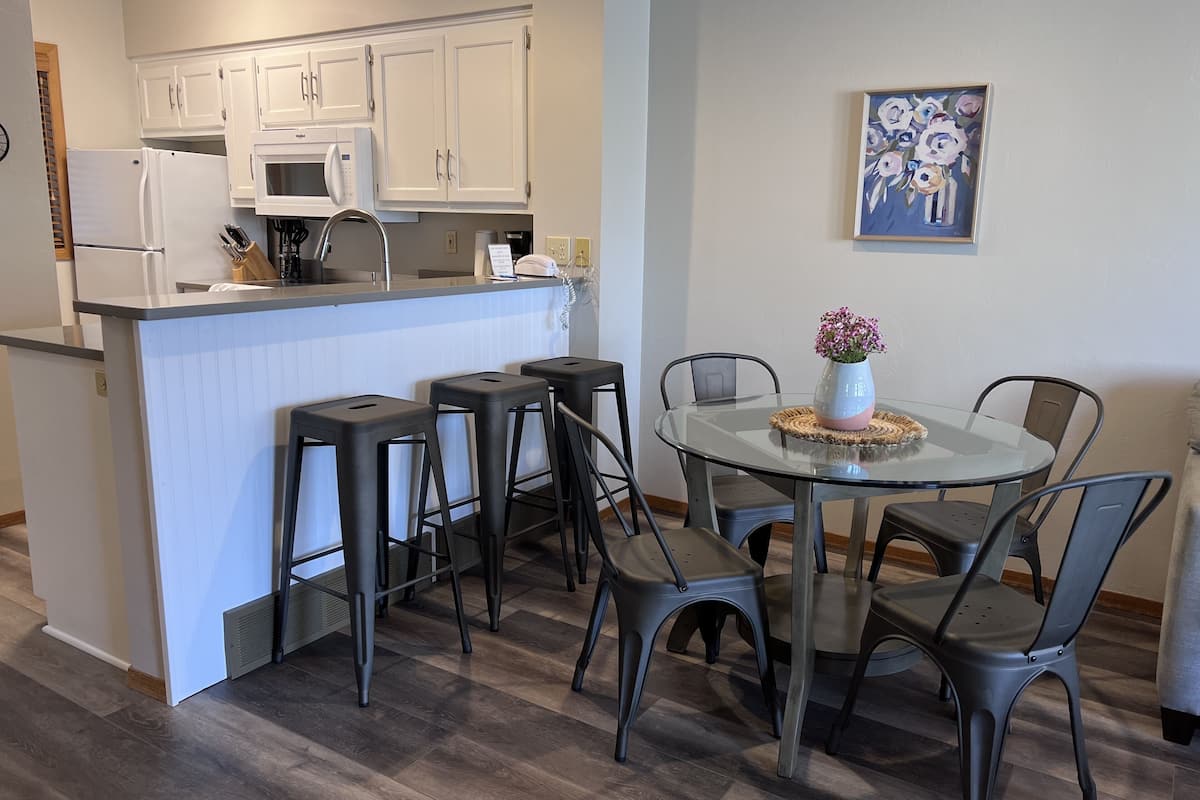 A modern kitchen with white cabinets, a microwave, and a coffee machine. There is a dining area with a glass table and four chairs. A vase with flowers is on the table.