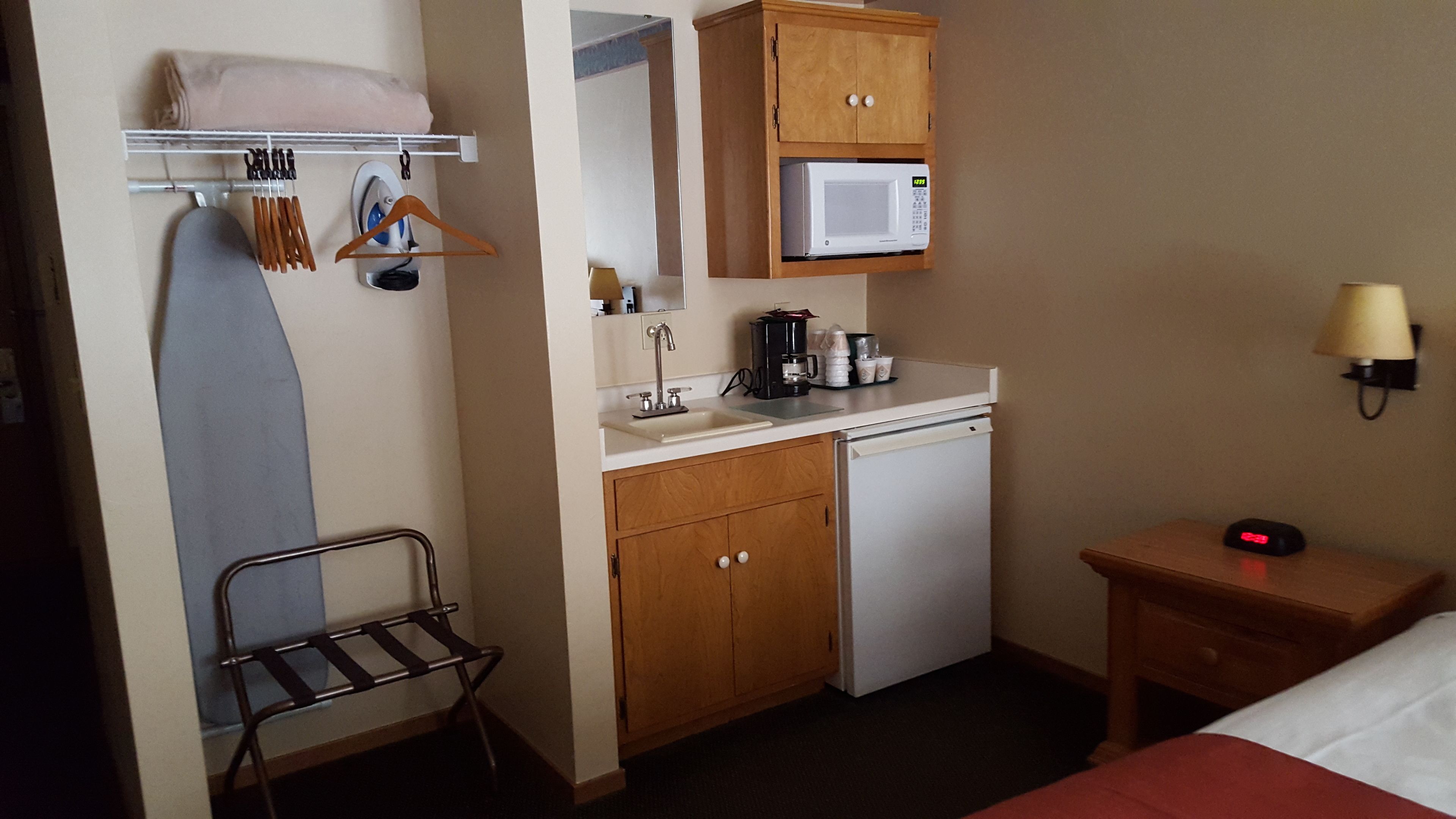 A compact hotel room with a kitchenette, a small ironing board, a microwave, a coffee maker, and a bedside table with a lamp.