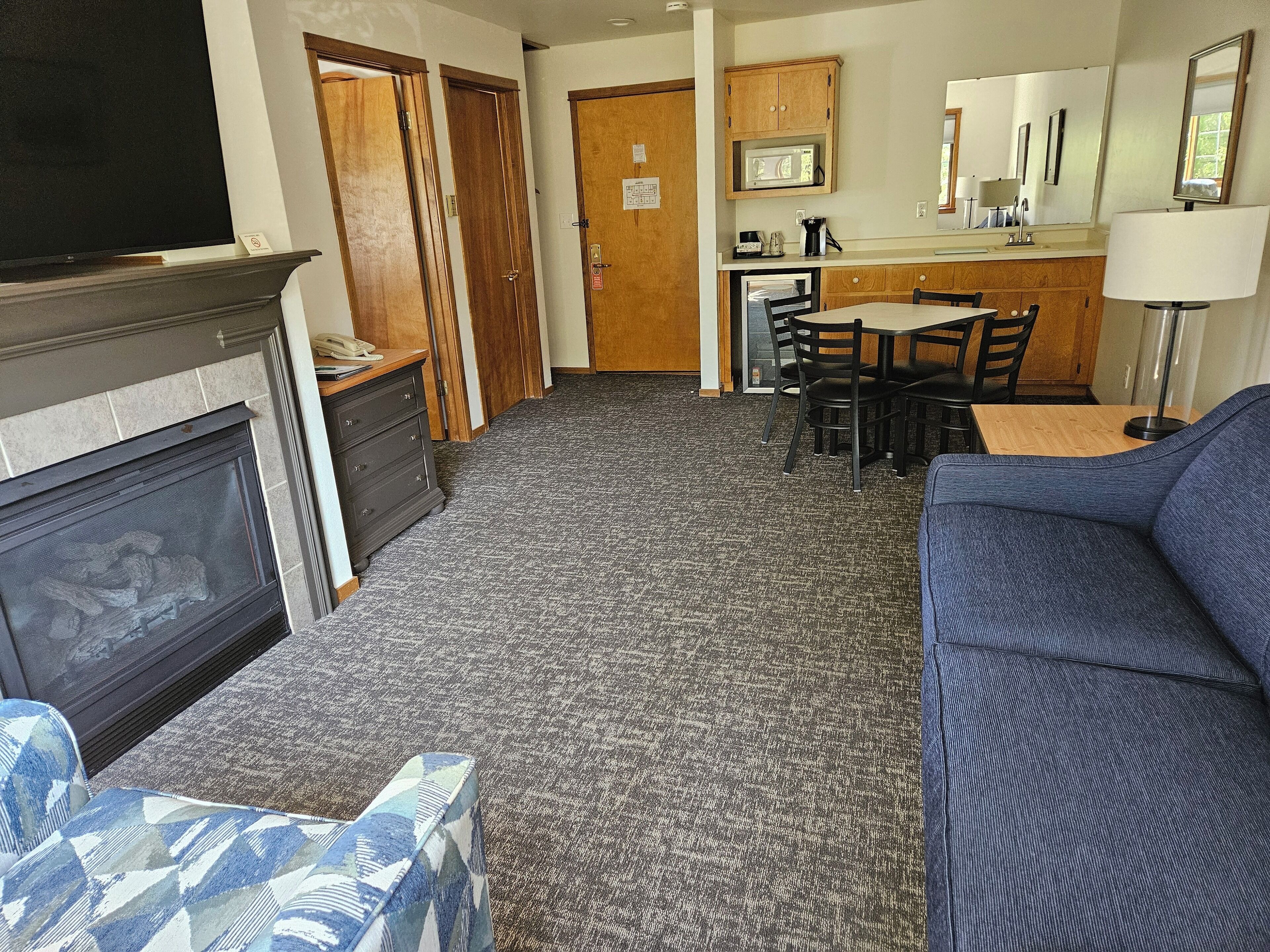 A hotel room with a fireplace, a sofa, a dining area with a table and chairs, and a kitchen area with a microwave and cabinets.