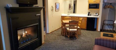 A living room with a fireplace, a television, a dining area with chairs, a kitchen with a microwave, and a bed.
