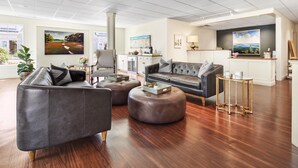 A spacious living room with a grey sofa, ottomans, and a coffee table. There are paintings on the walls and a kitchen area in the background.