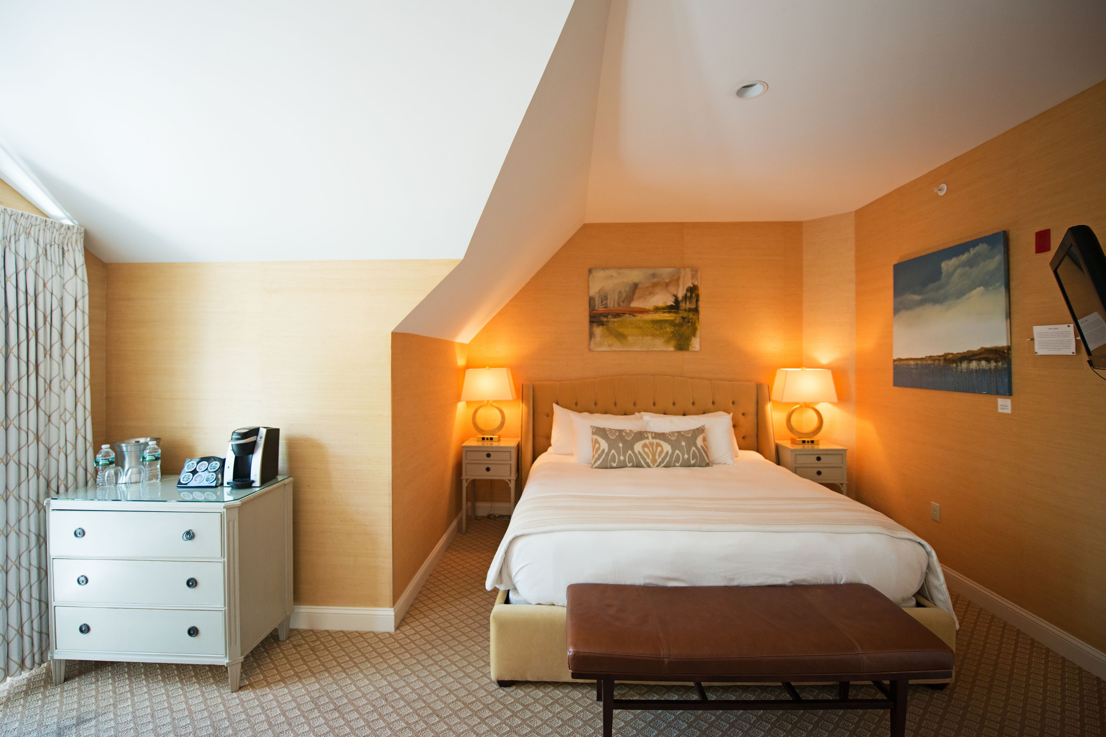 Una habitación de hotel con cama, mesitas de noche, un banco y obras de arte en las paredes.