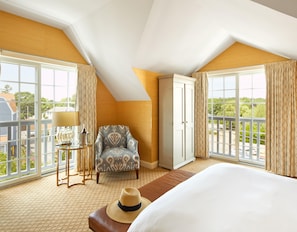Habitación de hotel con una cama grande, una silla, un armario, una mesita con lámpara y ventanas amplias con cortinas.