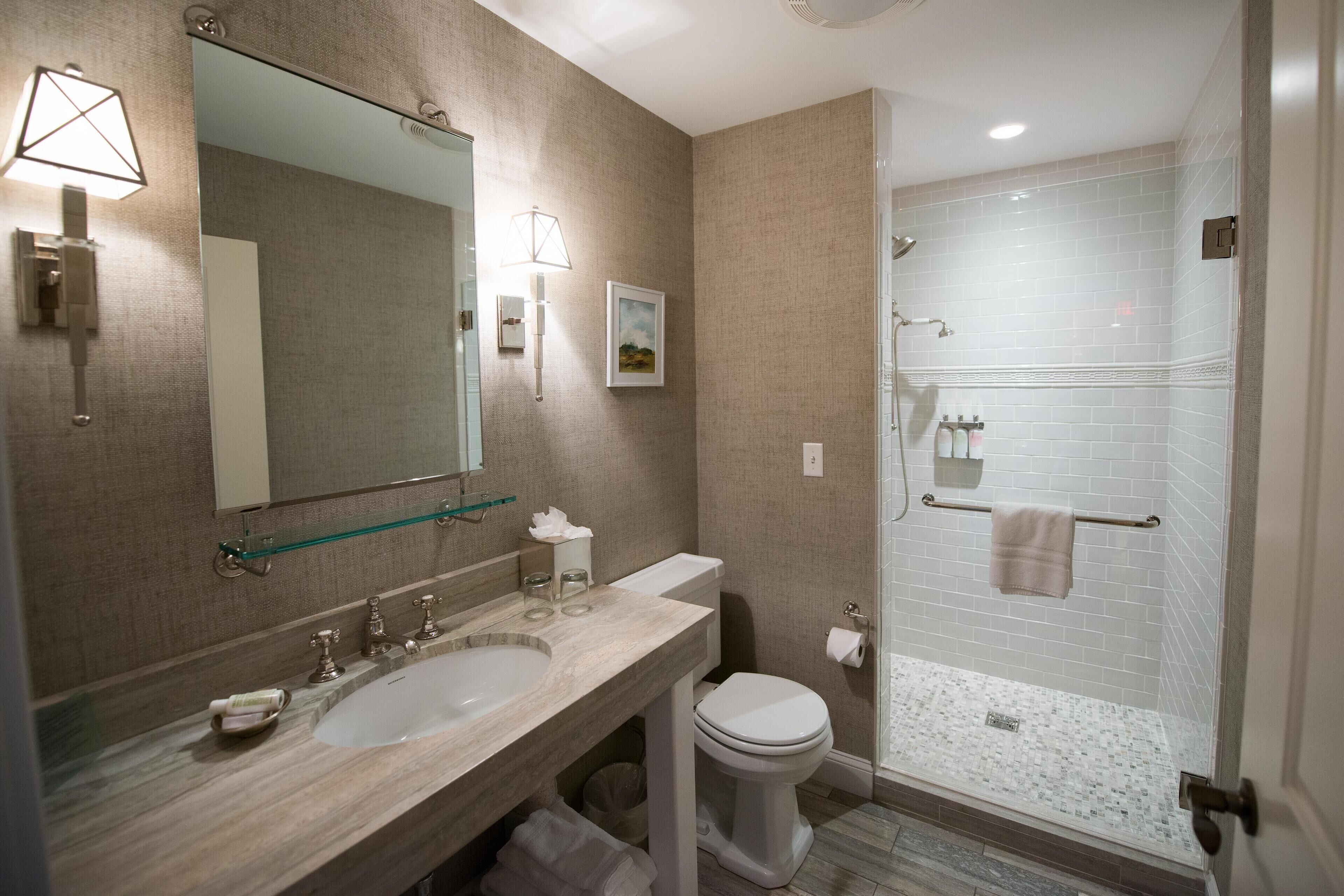 A modern bathroom with a large mirror, two sinks, a toilet, and a walk-in shower.