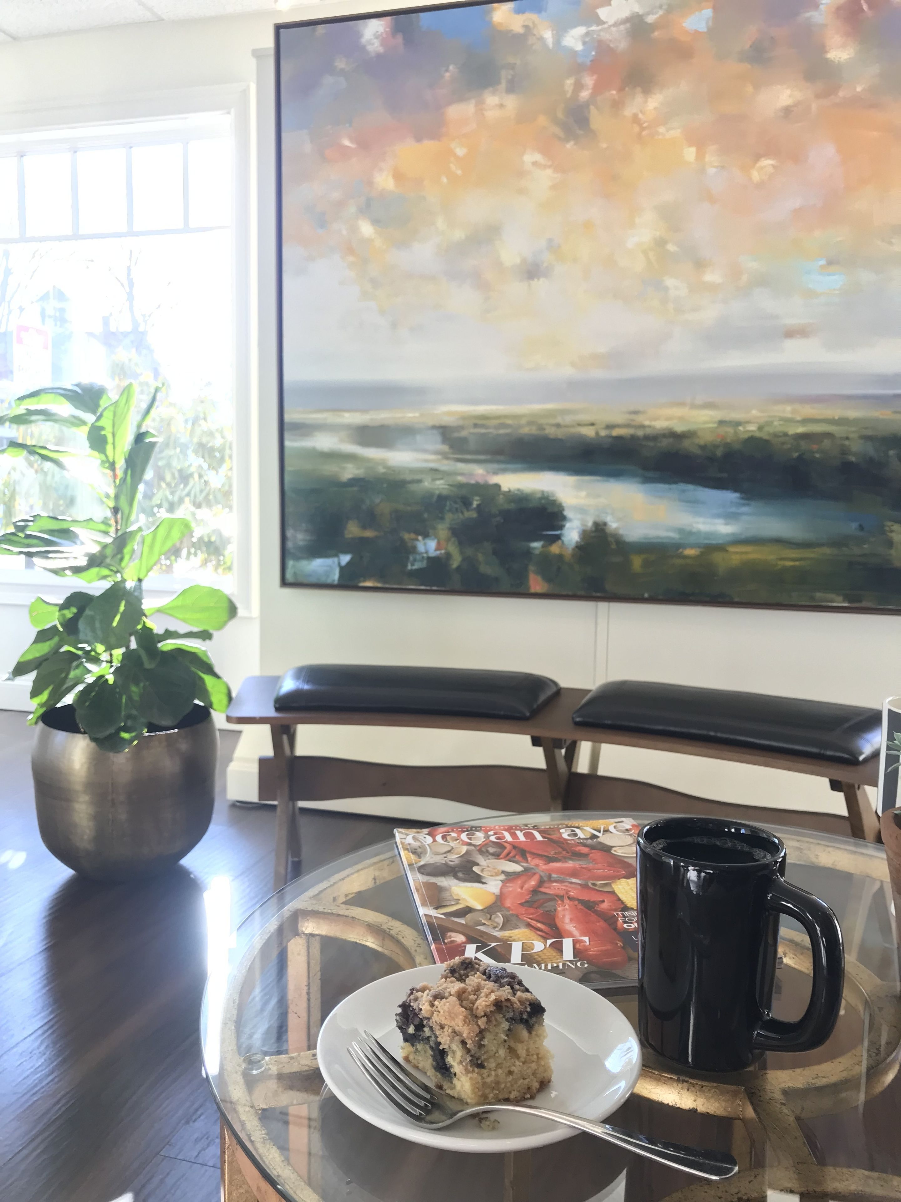 A dining table with a plate of food, a black mug, and a magazine. There is a potted plant and a bench in the background.