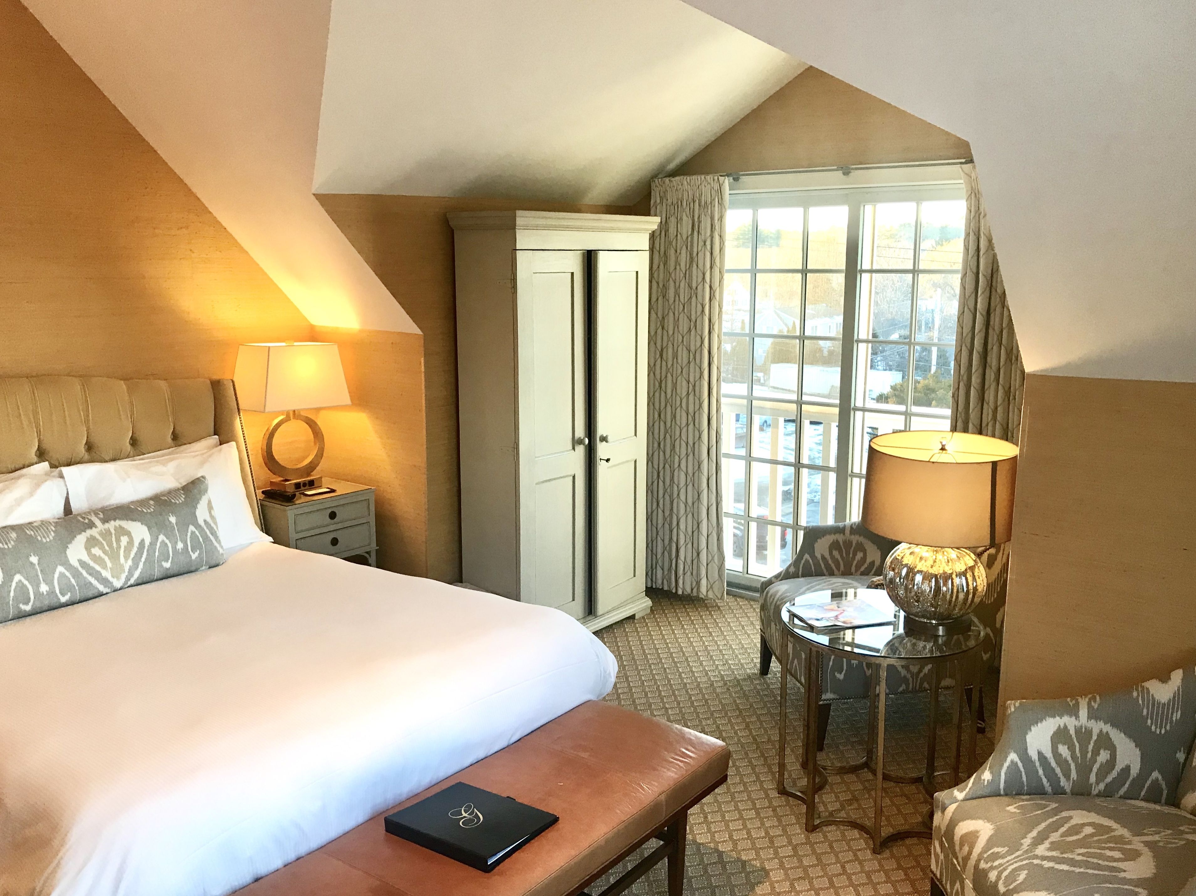 Habitación de hotel con una cama grande, una mesita de noche con lámpara, una pequeña mesa con lámpara, una silla y vistas al exterior a través de una ventana.