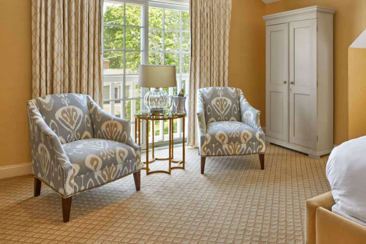 A room with two patterned armchairs, a small side table with a lamp, a large window with curtains, and a white wardrobe.