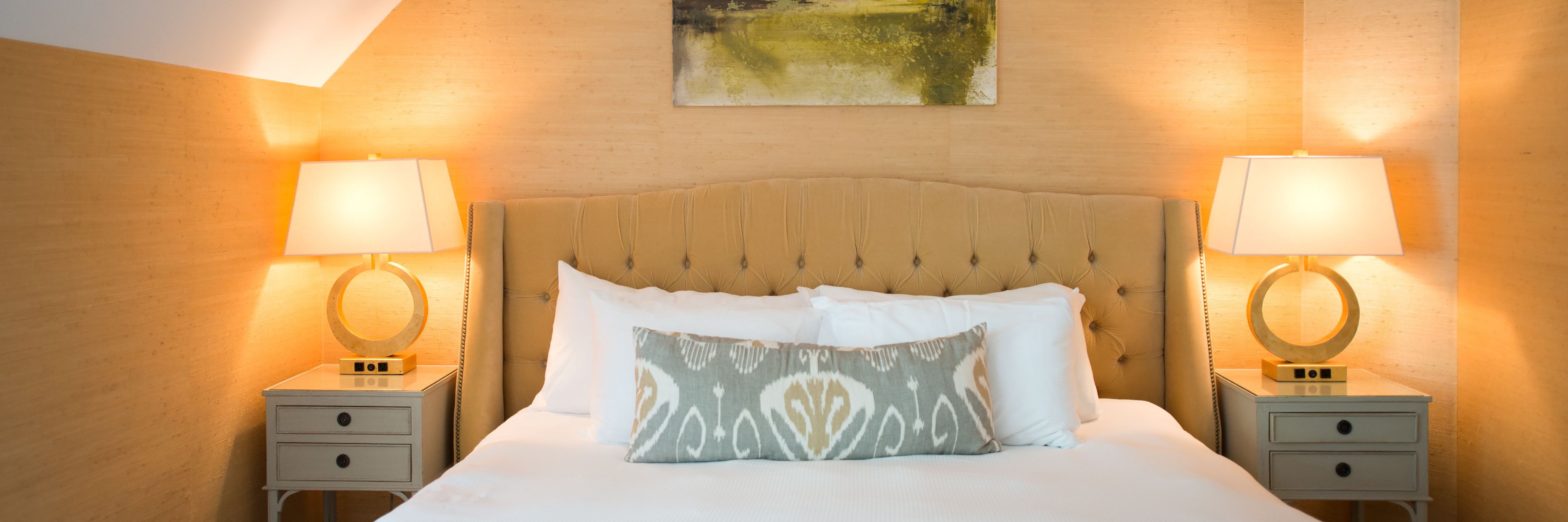 A neatly made bed with a tufted headboard, two bedside tables with lamps, and a painting on the wall.