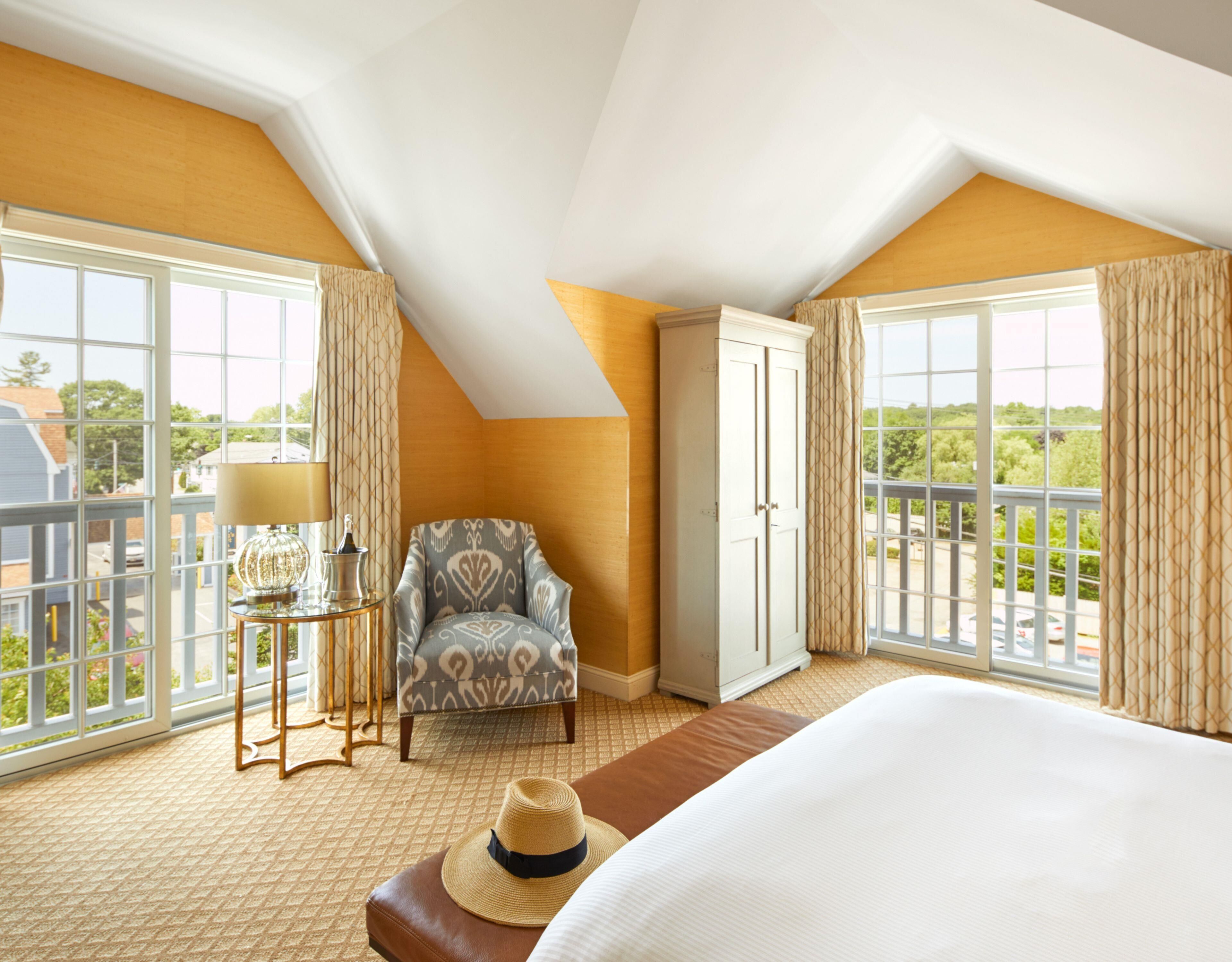 A hotel room with a large bed, a chair, a wardrobe, a small table with a lamp, and large windows with curtains.