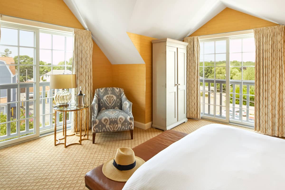A hotel room with a large bed, a chair, a wardrobe, a small table with a lamp, and large windows with curtains.