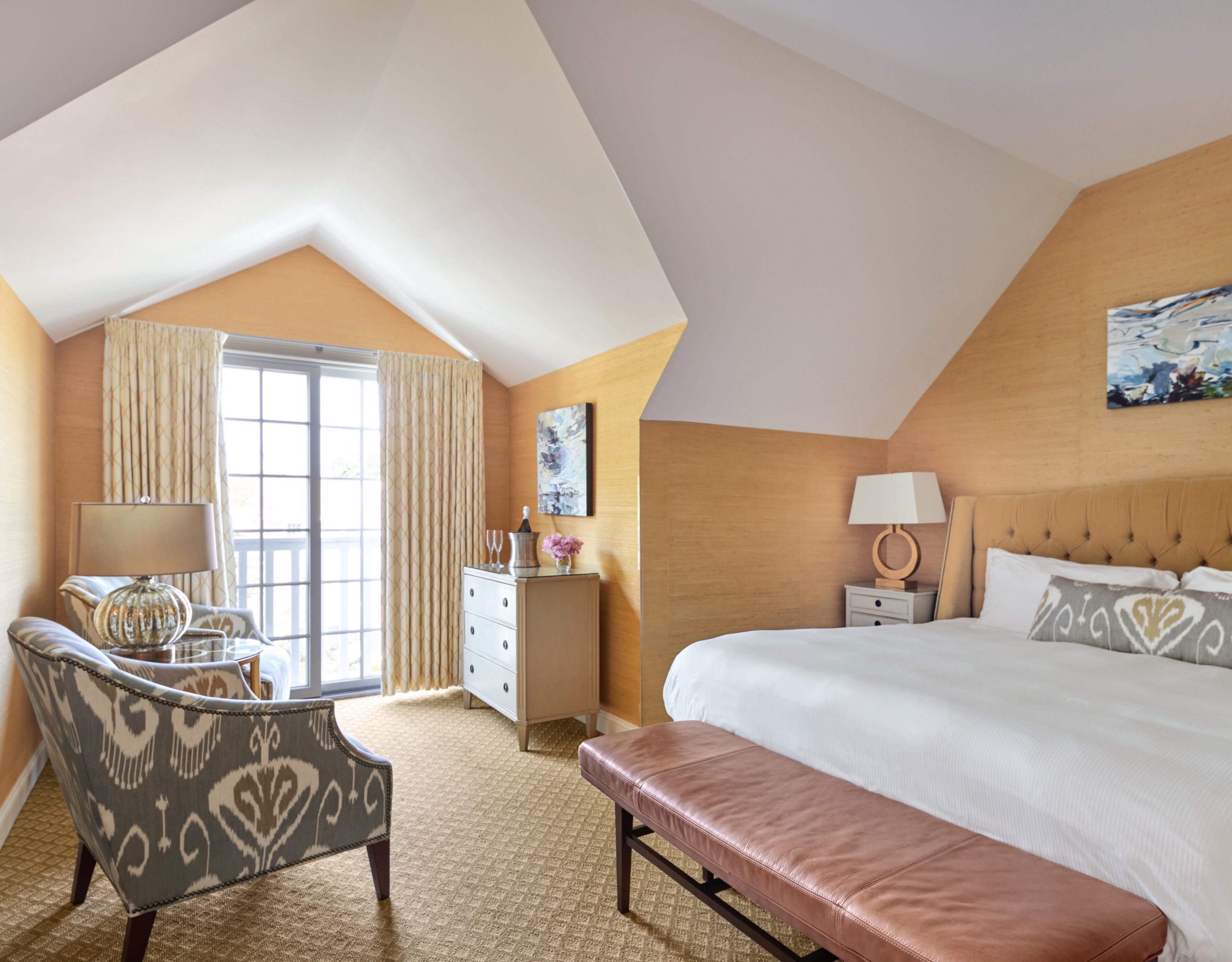 A hotel room with a large bed, a desk, a chair, a bench, a dresser, and a window with curtains.
