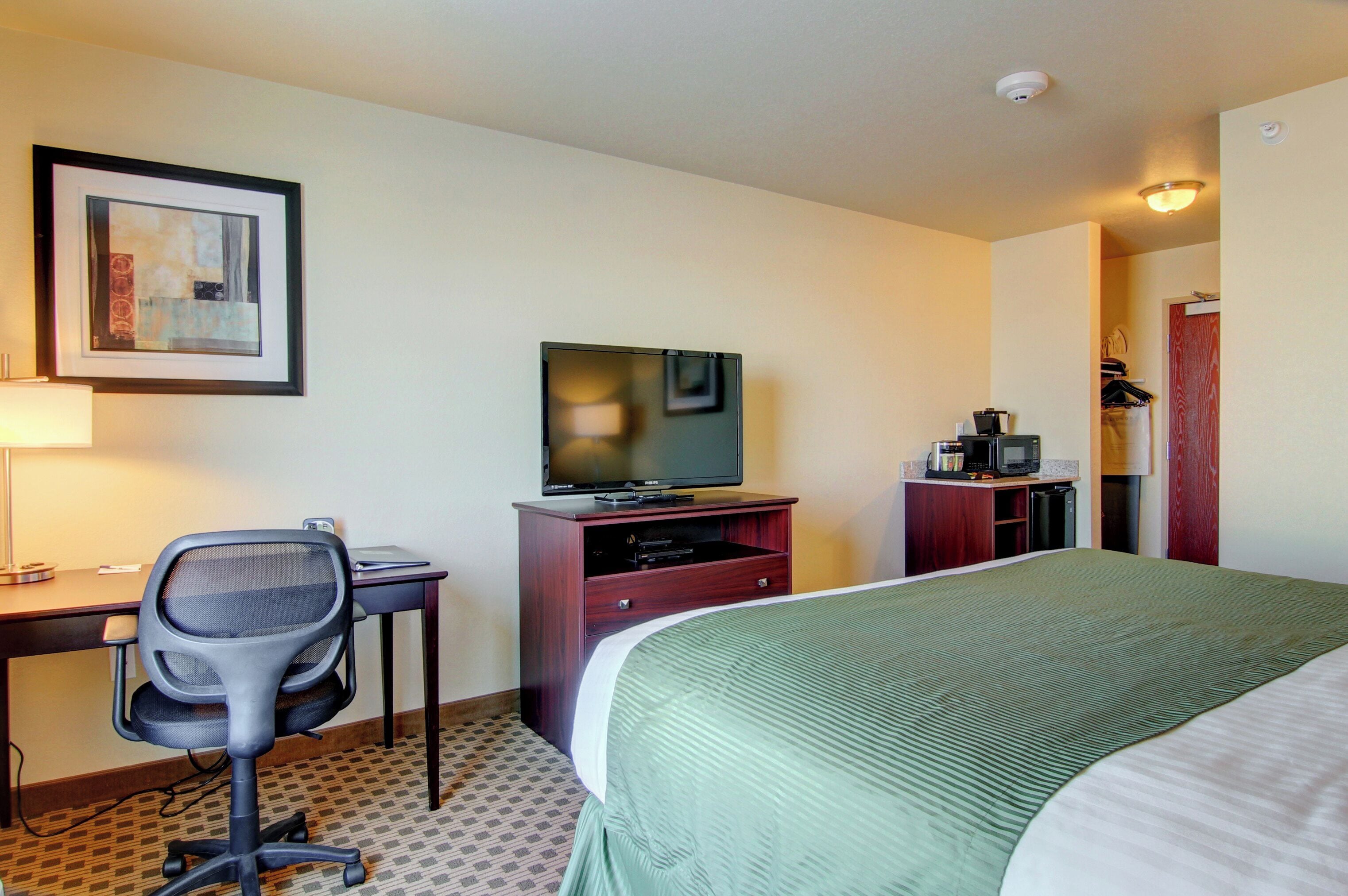 A hotel room with a bed, a desk with a chair, a television, a small kitchenette, and a framed picture on the wall.