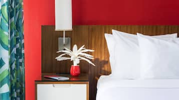 A hotel room with a red wall, a wooden headboard, white bedding, a bedside table with a lamp, a red vase with a plant, and a blue carpet.