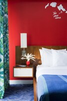 A hotel room with a red wall, a wooden headboard, white bedding, a bedside table with a lamp, a red vase with a plant, and a blue carpet.