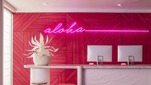 Uma área de recepção moderna com um letreiro de neon 'aloha', dois banquetes, um vaso de planta e uma TV de tela plana fixada na parede.