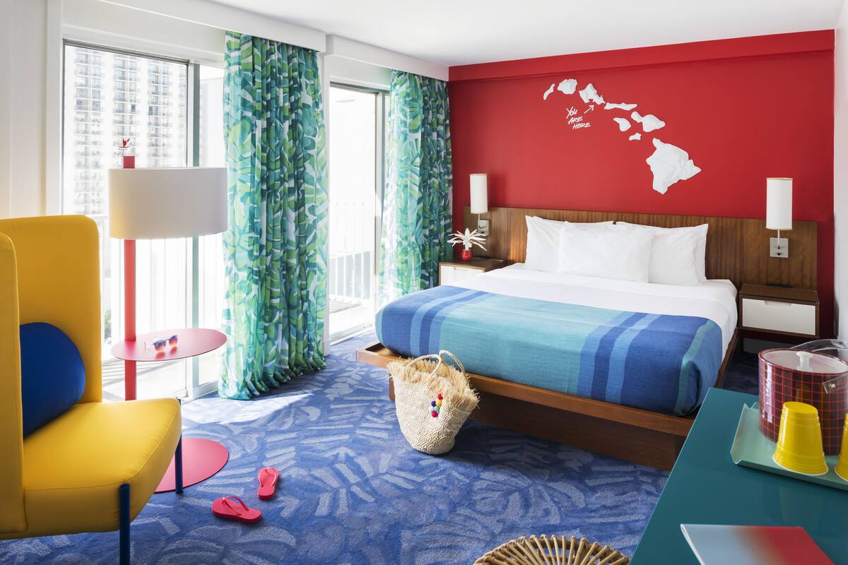 A hotel room with a bed, a chair, a small table, a lamp, and a window with curtains.