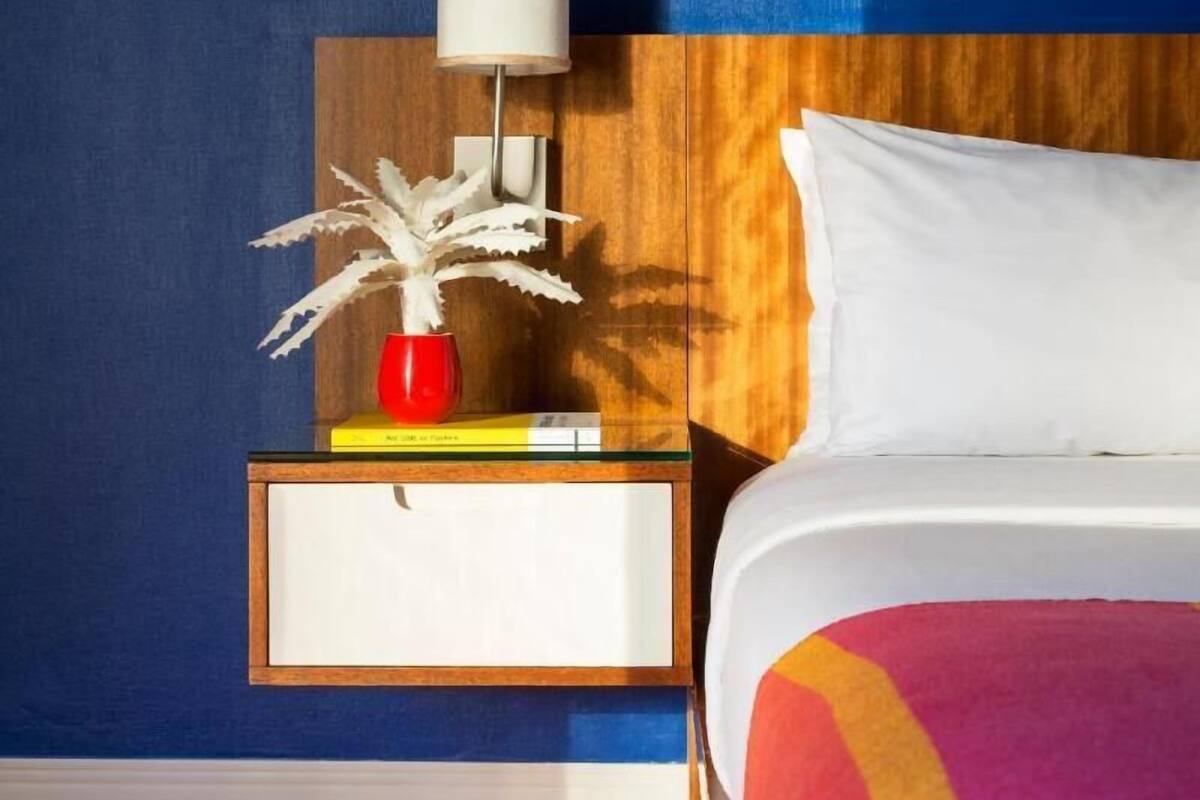 A hotel room with a bed, a nightstand with a lamp, a red vase, and a framed picture.