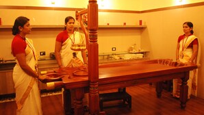 A room with a wooden table, a scale, and three individuals in traditional attire. - Country Spa Wellness Beach Resort (Thiruvananthapuram)