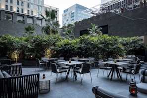 Uma área de estar ao ar livre com mesas e cadeiras, cercada por vegetação e edifícios modernos ao fundo.