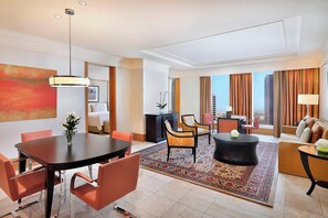 A modern hotel room with a dining area, a living room with a sofa and armchairs, a large window with curtains, and a view of buildings.