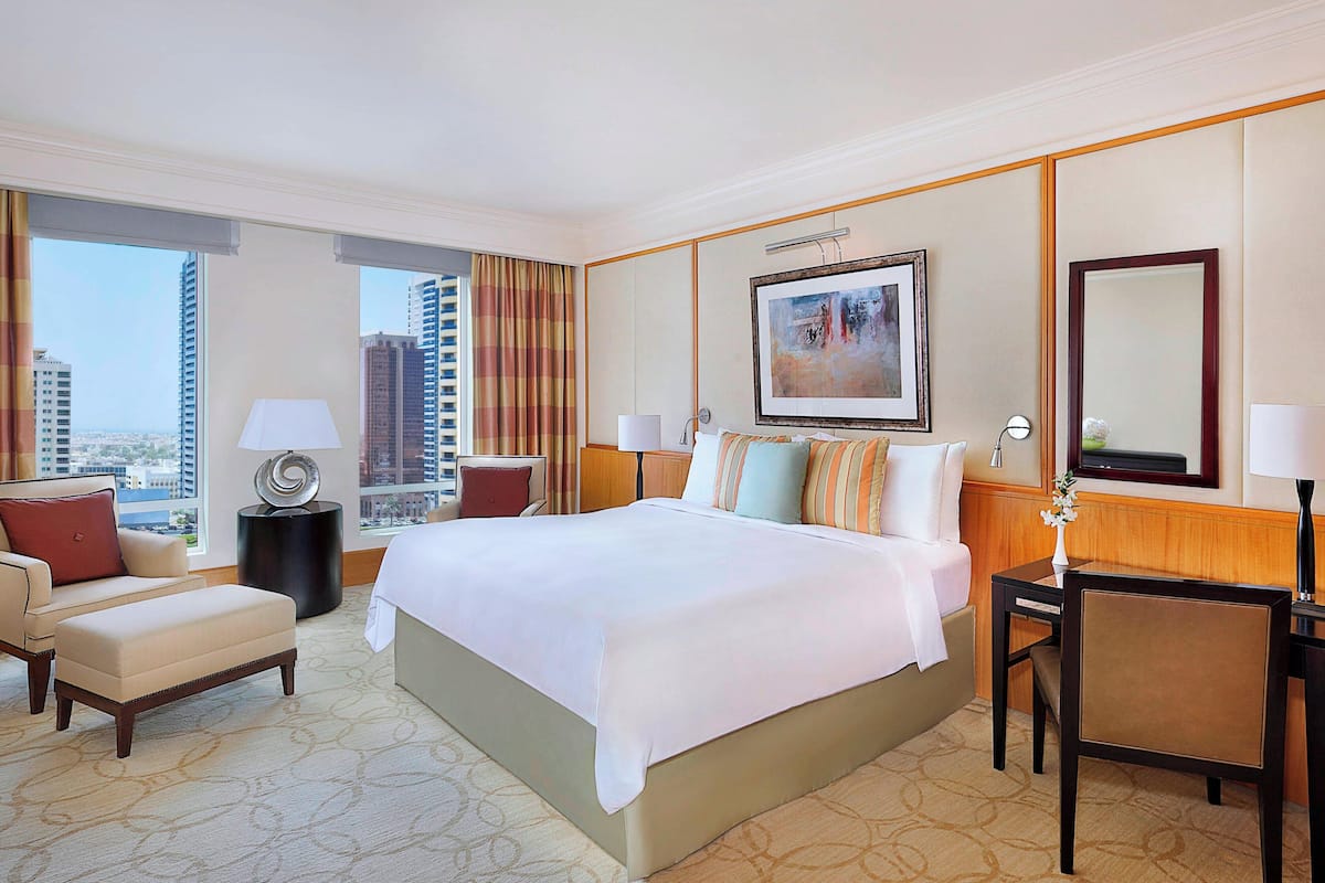 A hotel room with a large bed, a desk, a chair, a lamp, a painting, and a view of the city.